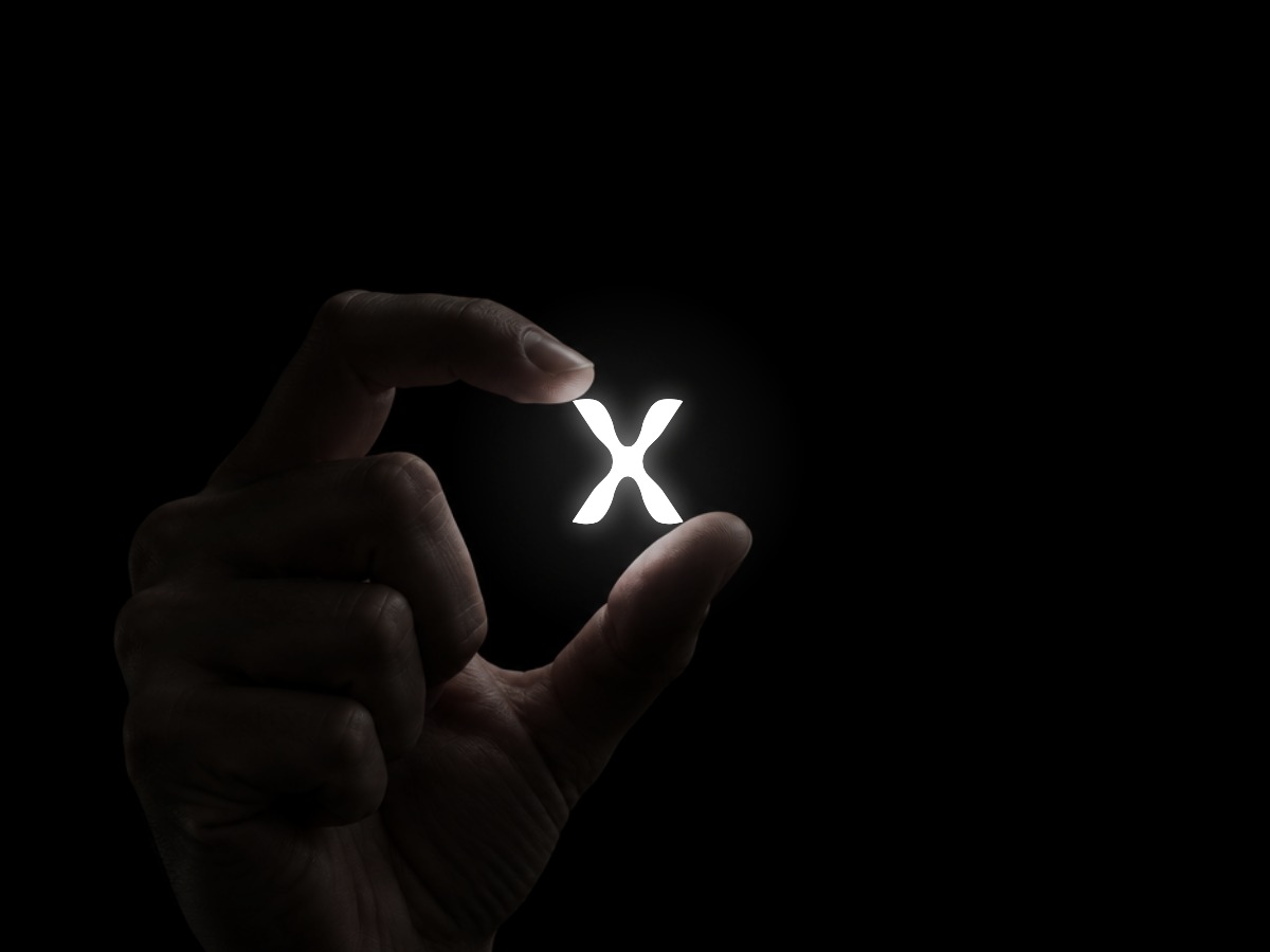 A dimly lit hand rising from below, holding the glowing Xctrl X with the index fingertip on its top-left corner and the thumb on its bottom-right corner